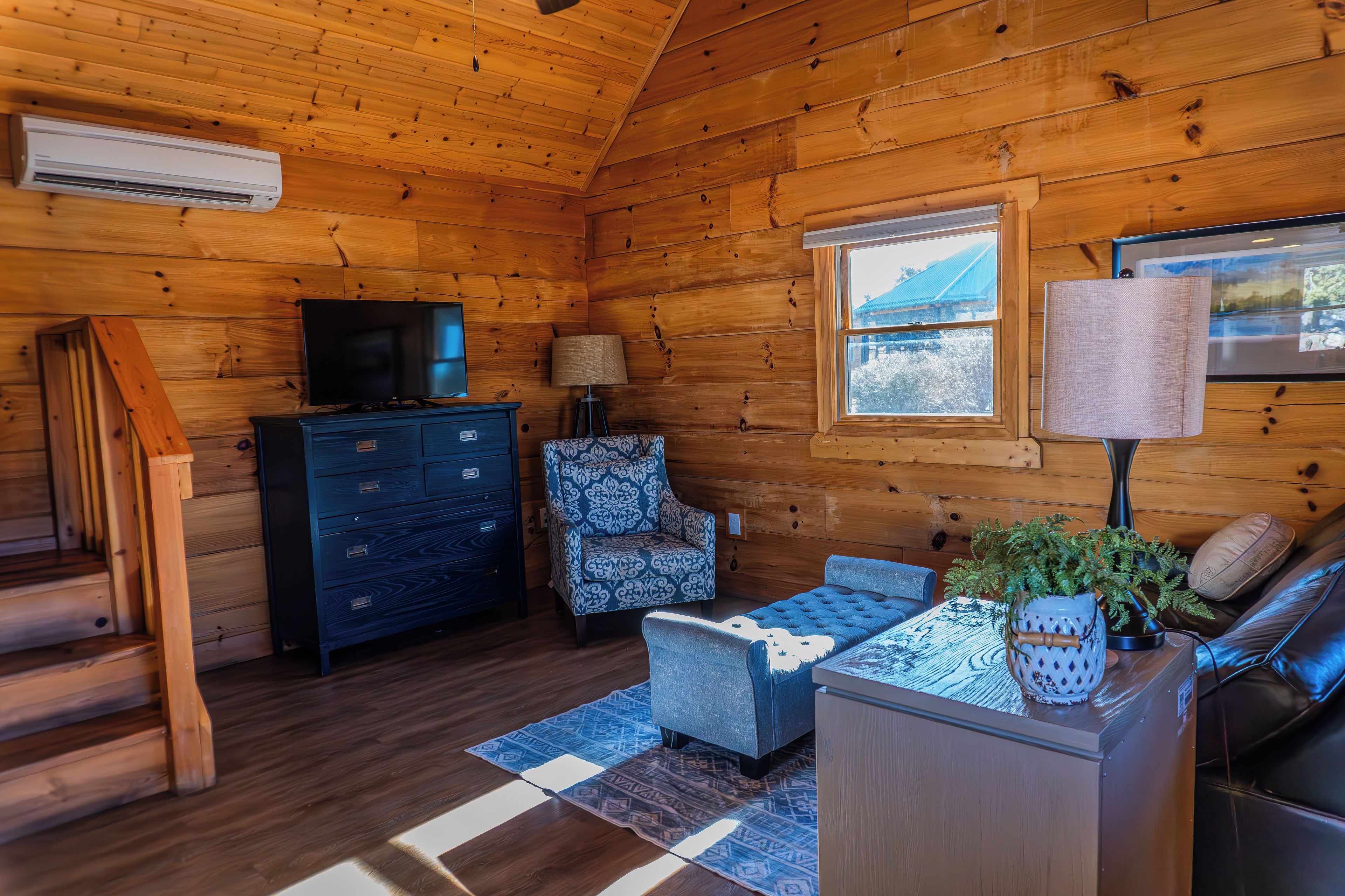 A log cabin interior with a television, a patterned armchair, a sofa, and a window with a view of a blue building.