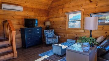 A log cabin interior with a television, a patterned armchair, a sofa, and a window with a view of a blue building.