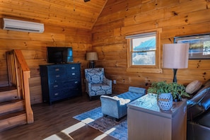 A log cabin interior with a television, a patterned armchair, a sofa, and a window with a view of a blue building.