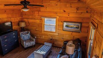 A log cabin interior with a ceiling fan, a television, a patterned armchair, a cushioned bench, a sofa, and a framed picture on the wall.