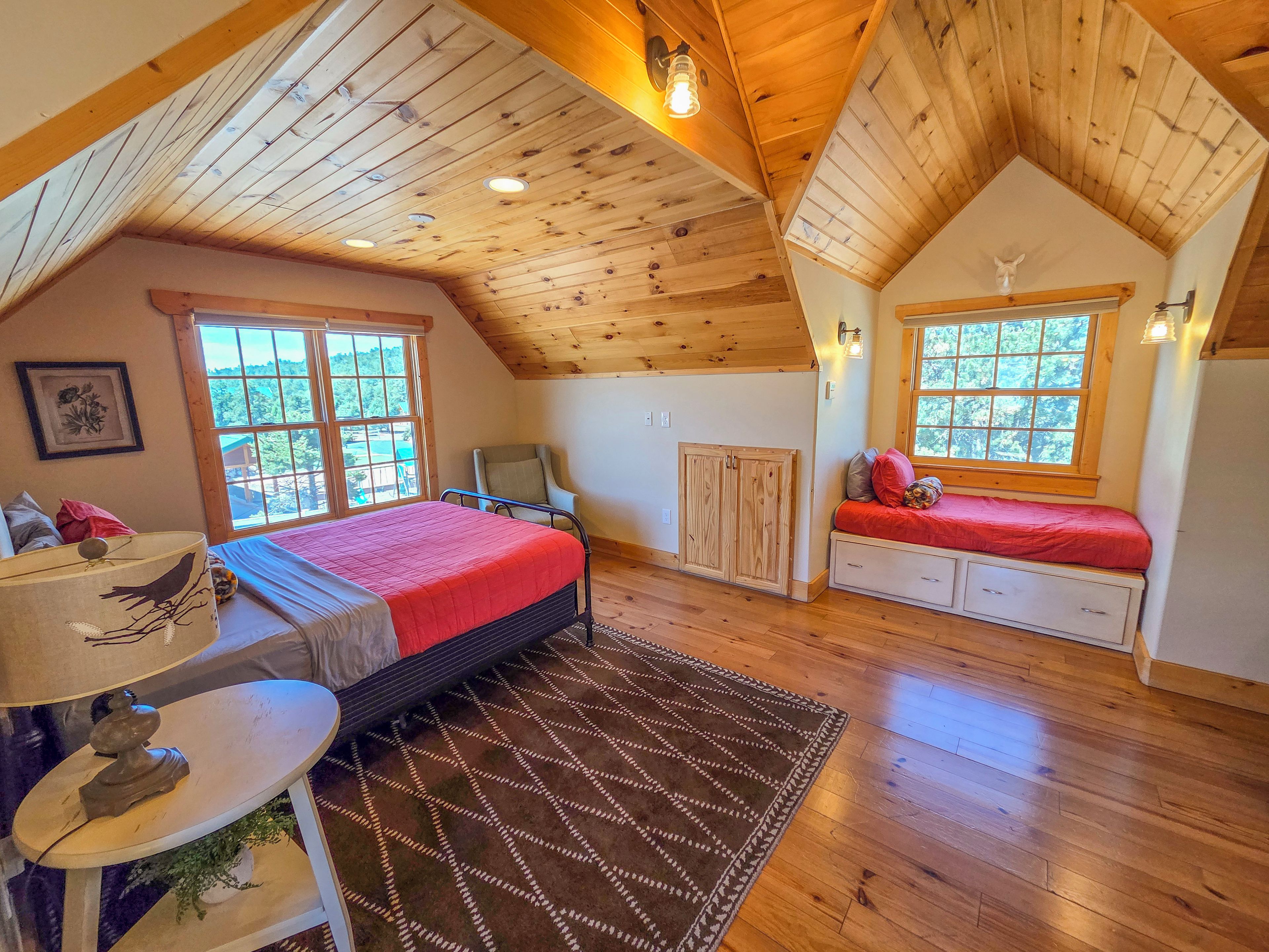 A bedroom with a sloped ceiling, a bed, a desk, a chair, and a window with a view of trees.