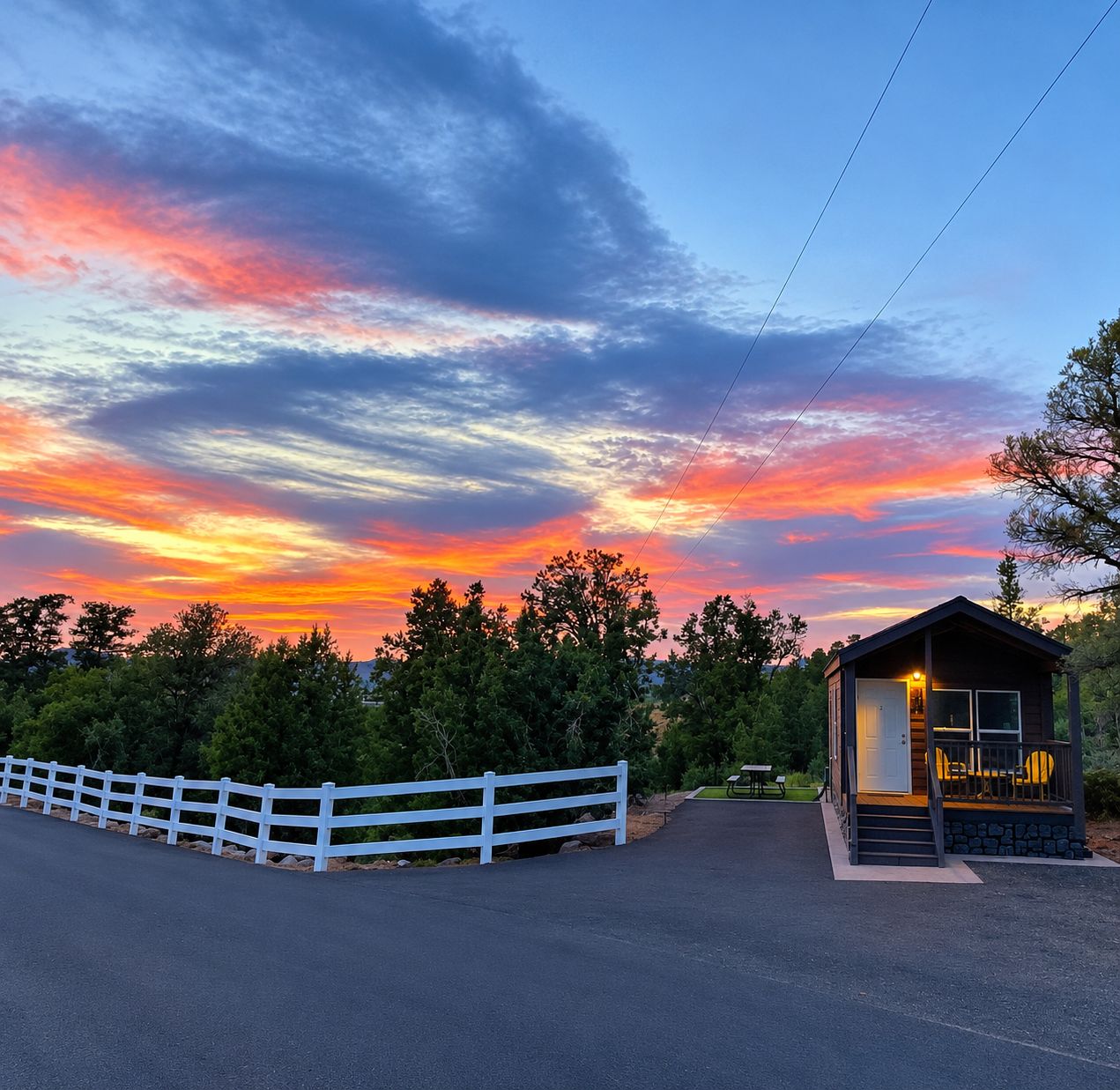 A small cabin with a porch, surrounded by a white fence, set against a sunset sky.