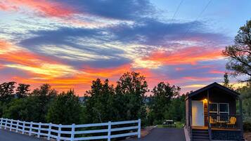 A small cabin with a porch, surrounded by a white fence, set against a sunset sky.