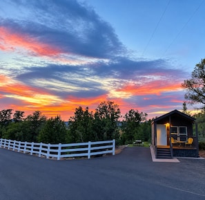 A small cabin with a porch, surrounded by a white fence, set against a sunset sky. - Holmstead Ranch Resort (Veyo)