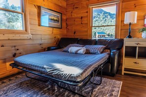 A wooden cabin interior with a bed, a nightstand, a lamp, and a window with a view of trees and mountains.