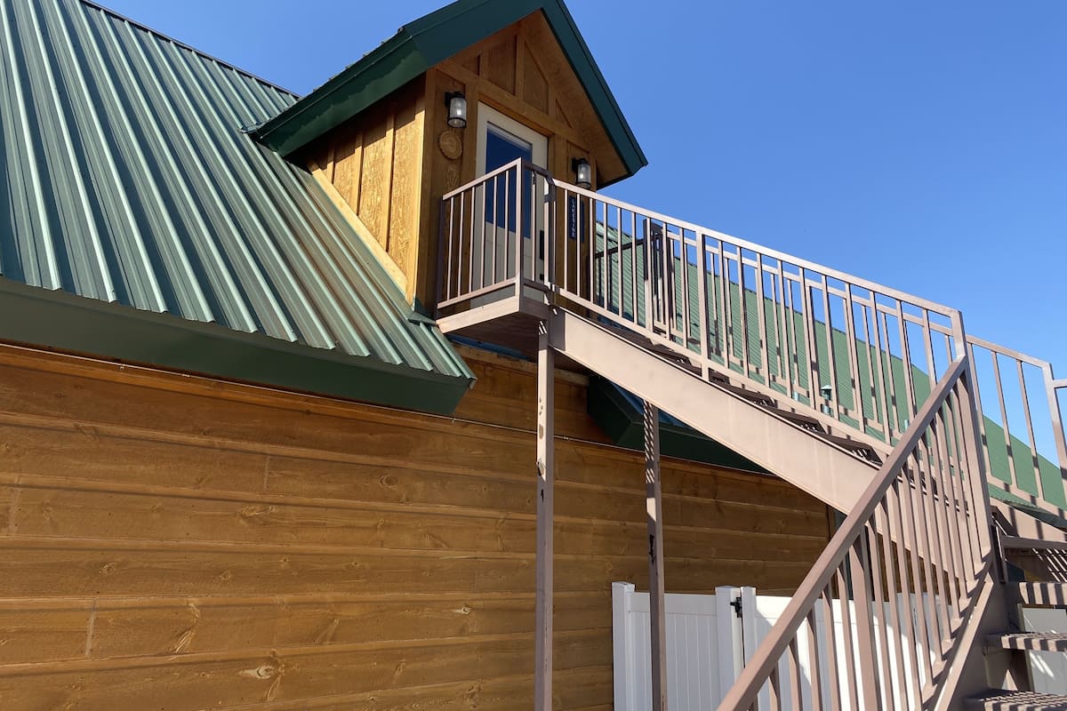 A wooden building with a green roof, a balcony with a metal railing, and a staircase leading to a door.