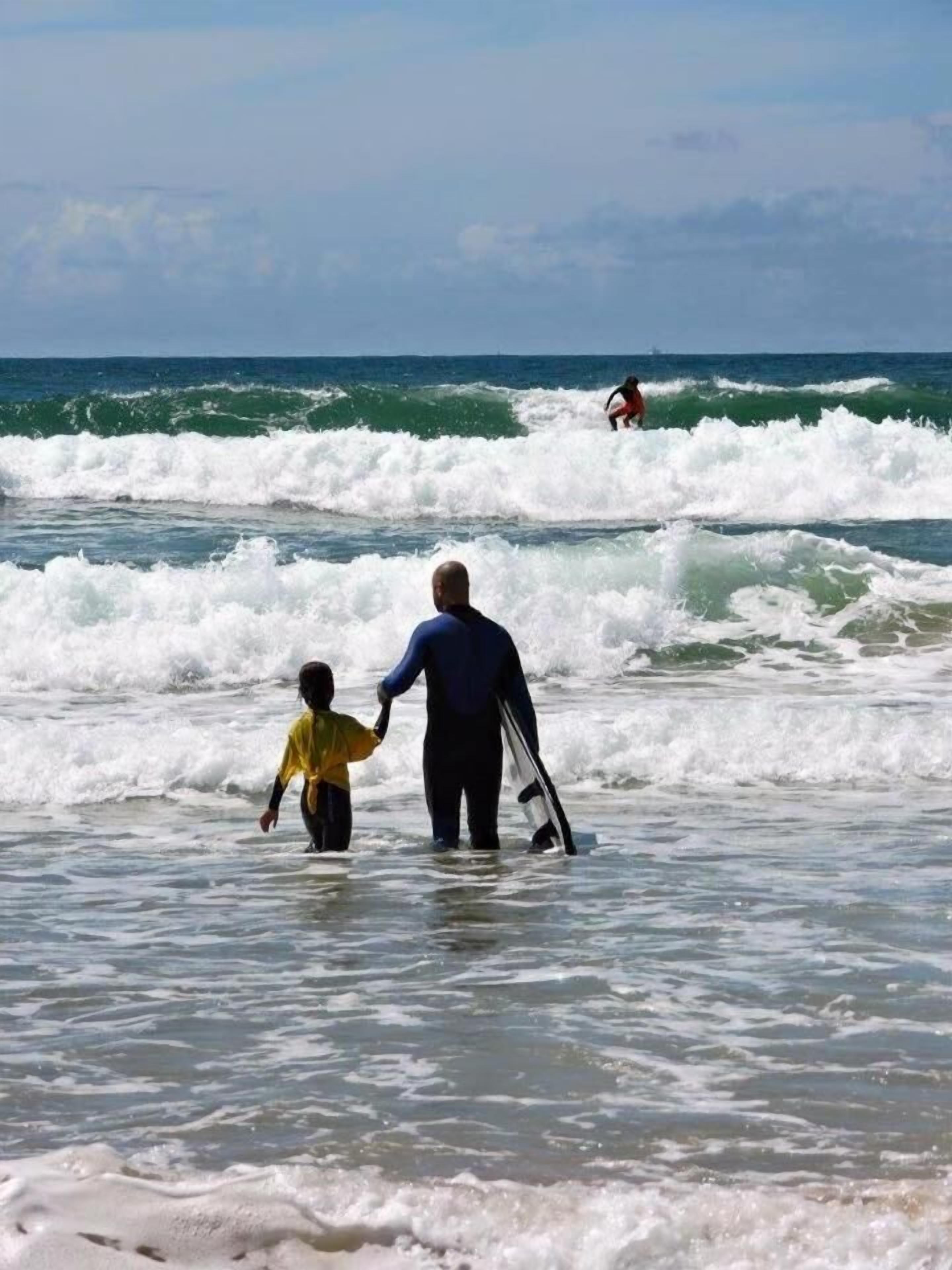 A beach scene with waves, a person holding a child, and another person surfing in the background.