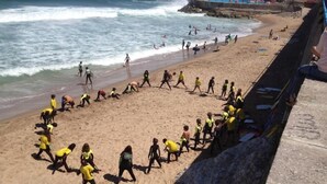 Una playa con gente usando camisas amarillas, una costa rocosa y un edificio al fondo.
