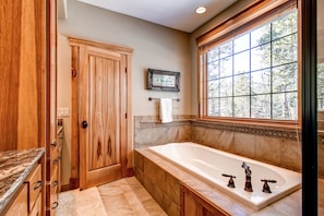 A bathroom with a large bathtub, wooden cabinetry, a window with a view of trees, and a framed picture on the wall.