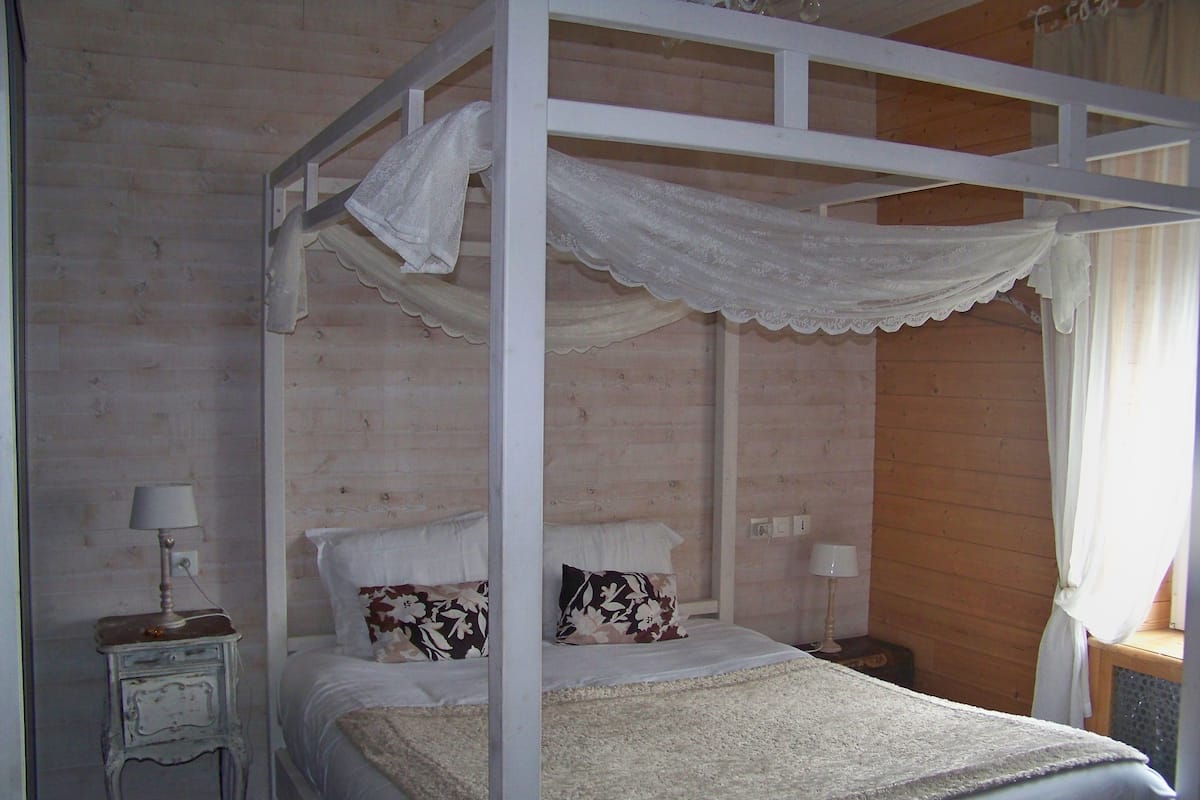 A four-poster bed with white bedding and lace canopy, a nightstand with a lamp, and a floral pillow.