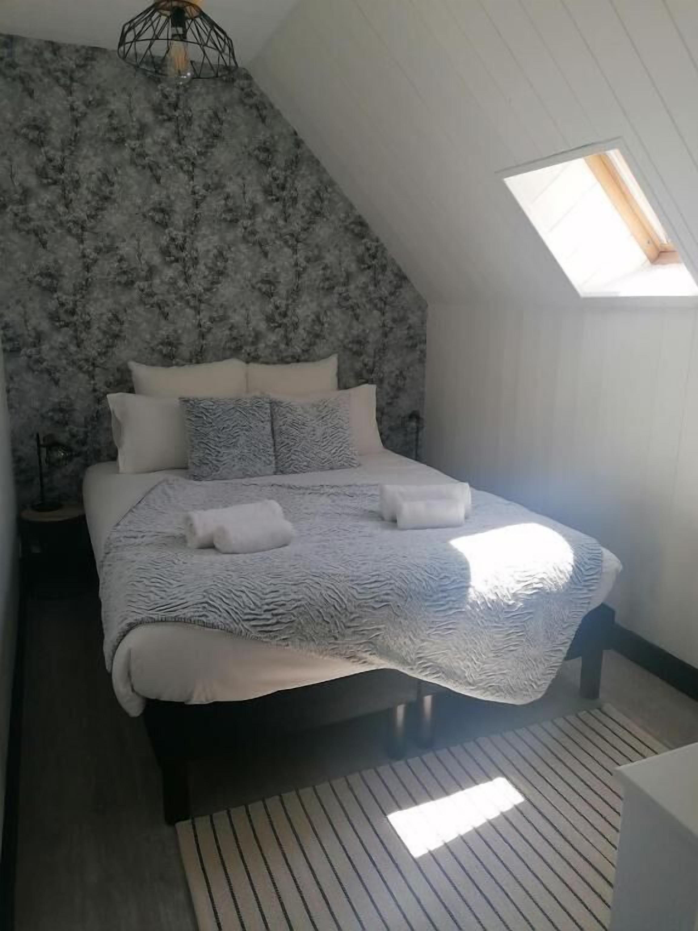 A bedroom with a sloped ceiling, a bed with a patterned blanket, pillows, and a bedside lamp.