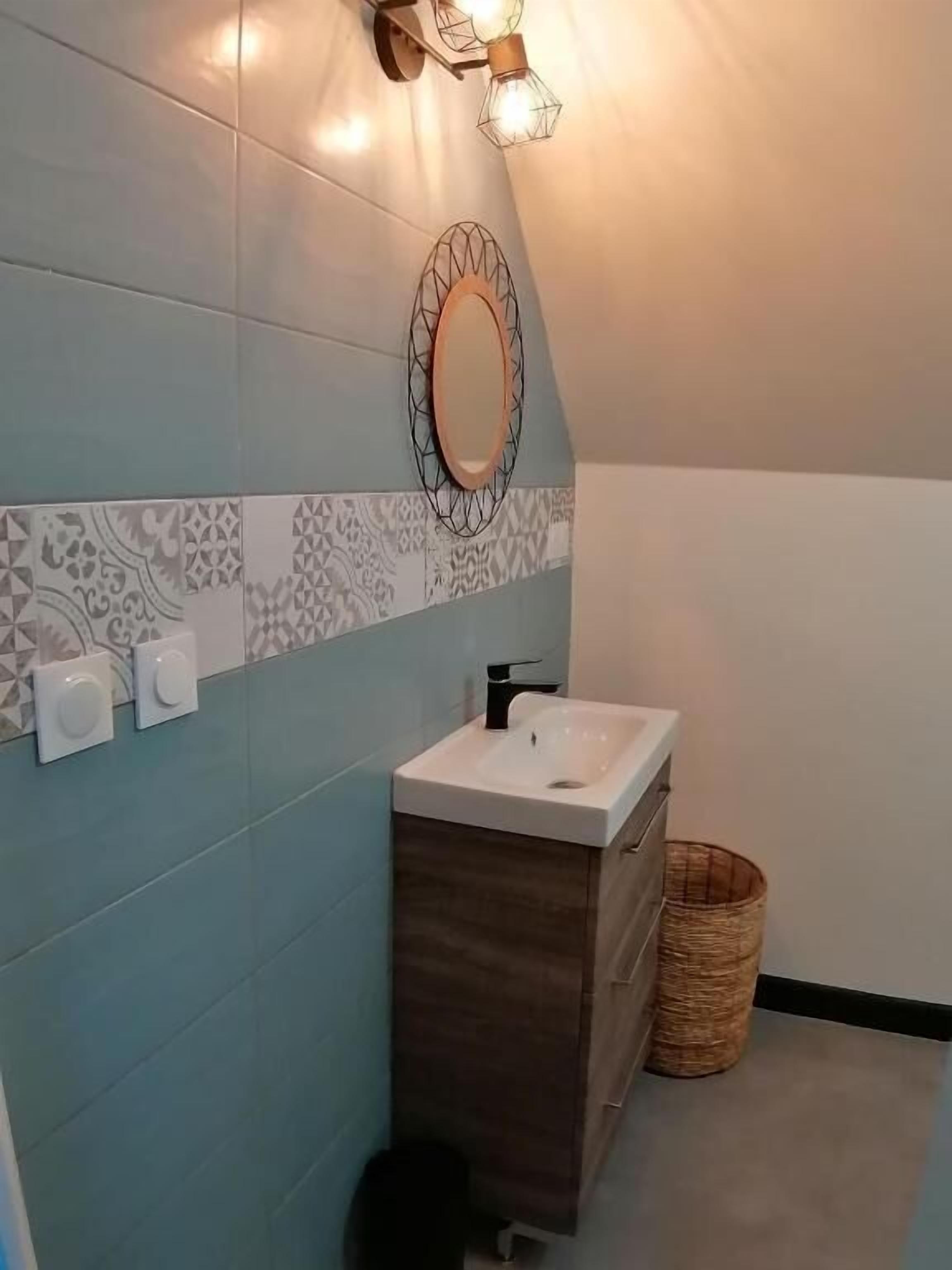 Un baño con pared de azulejos, un lavamanos, un espejo y una lámpara fijada a la pared.