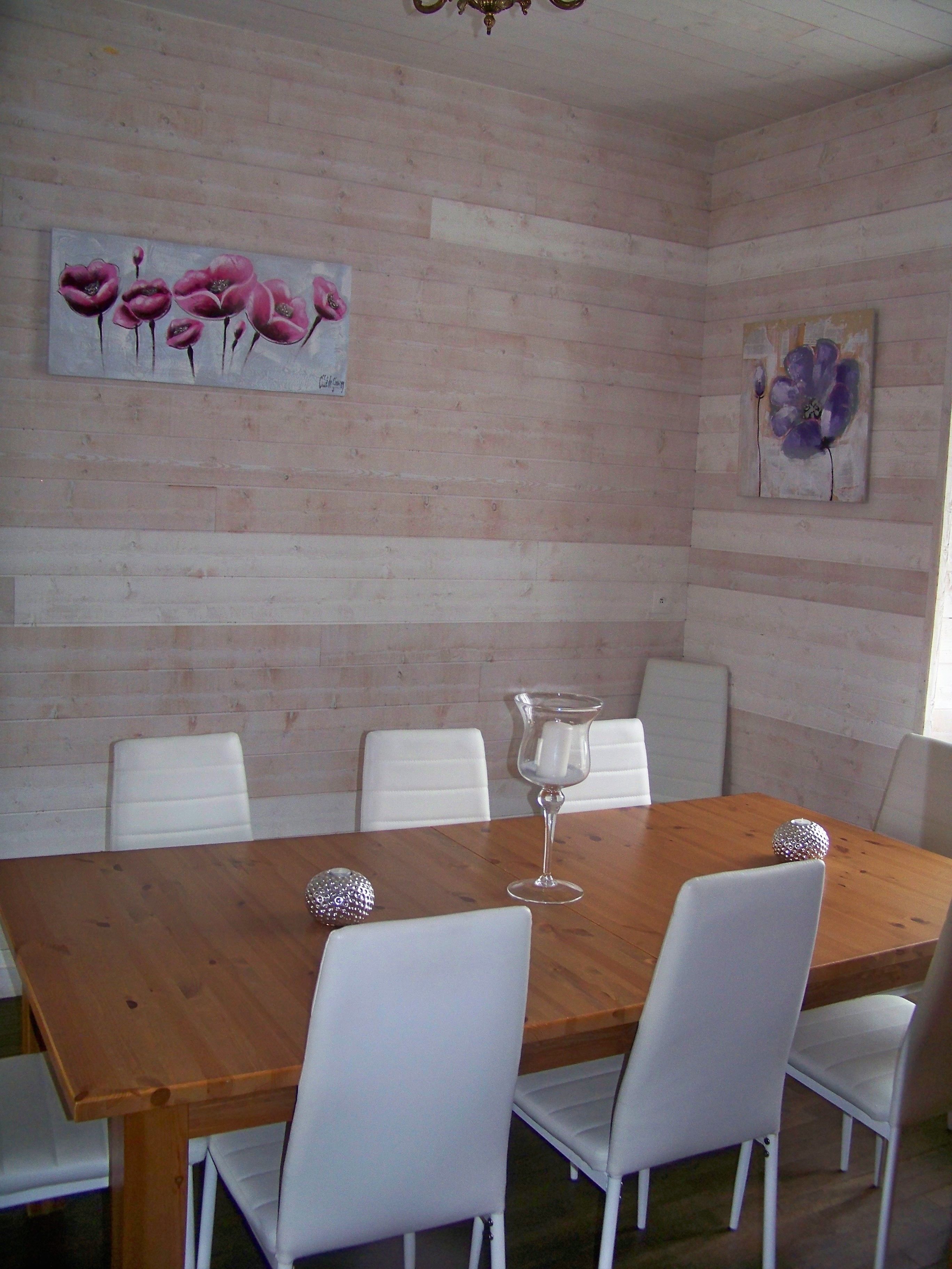 Uma sala de jantar com mesa de madeira e cadeiras brancas, um candelabro e duas pinturas florais nas paredes.