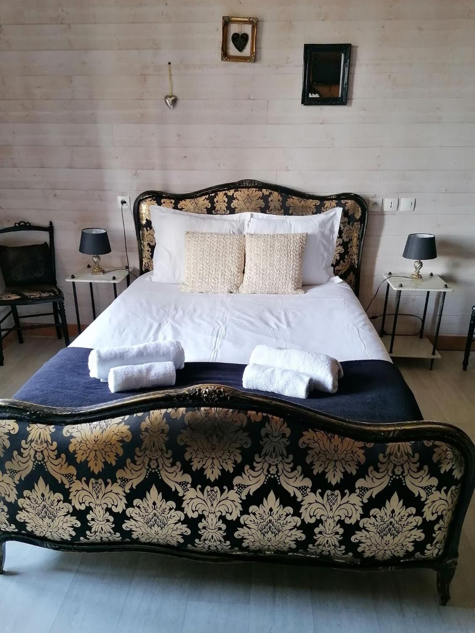 Una cama con un cabecero oscuro y decorativo, dos mesitas de noche con lámparas, y una pared con dos cuadros enmarcados.