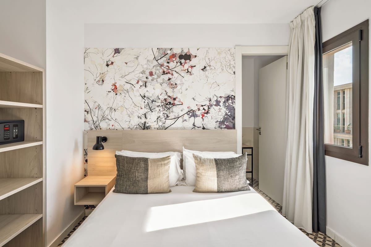 A modern hotel room with a large bed, a headboard with a floral mural, a bedside table with a lamp, and a view of the city through a window.