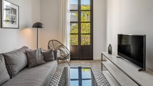 A modern living room with a sofa, a TV, a lamp, and a door leading to a balcony with a view of trees. - Aspasios Poblenou Apartments (Barcelona)