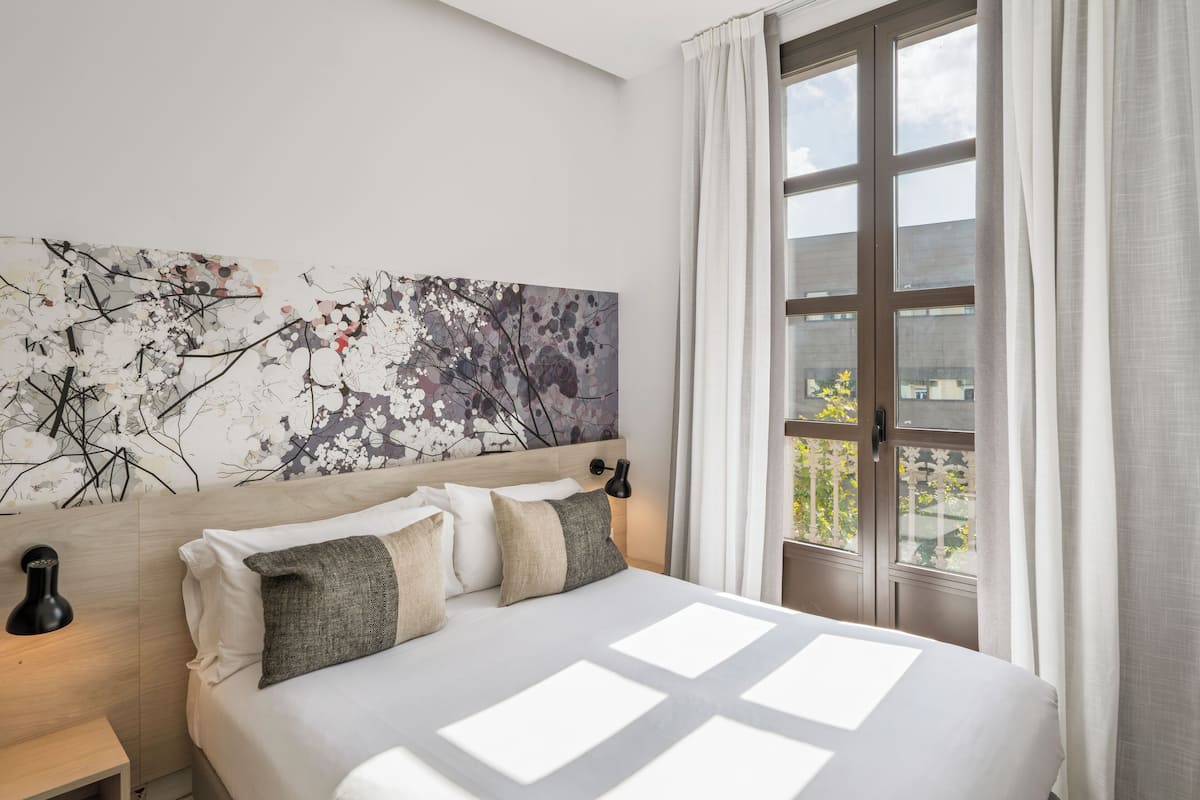 A hotel room with a large bed, a headboard with a floral artwork, a nightstand with a lamp, and a window with curtains.