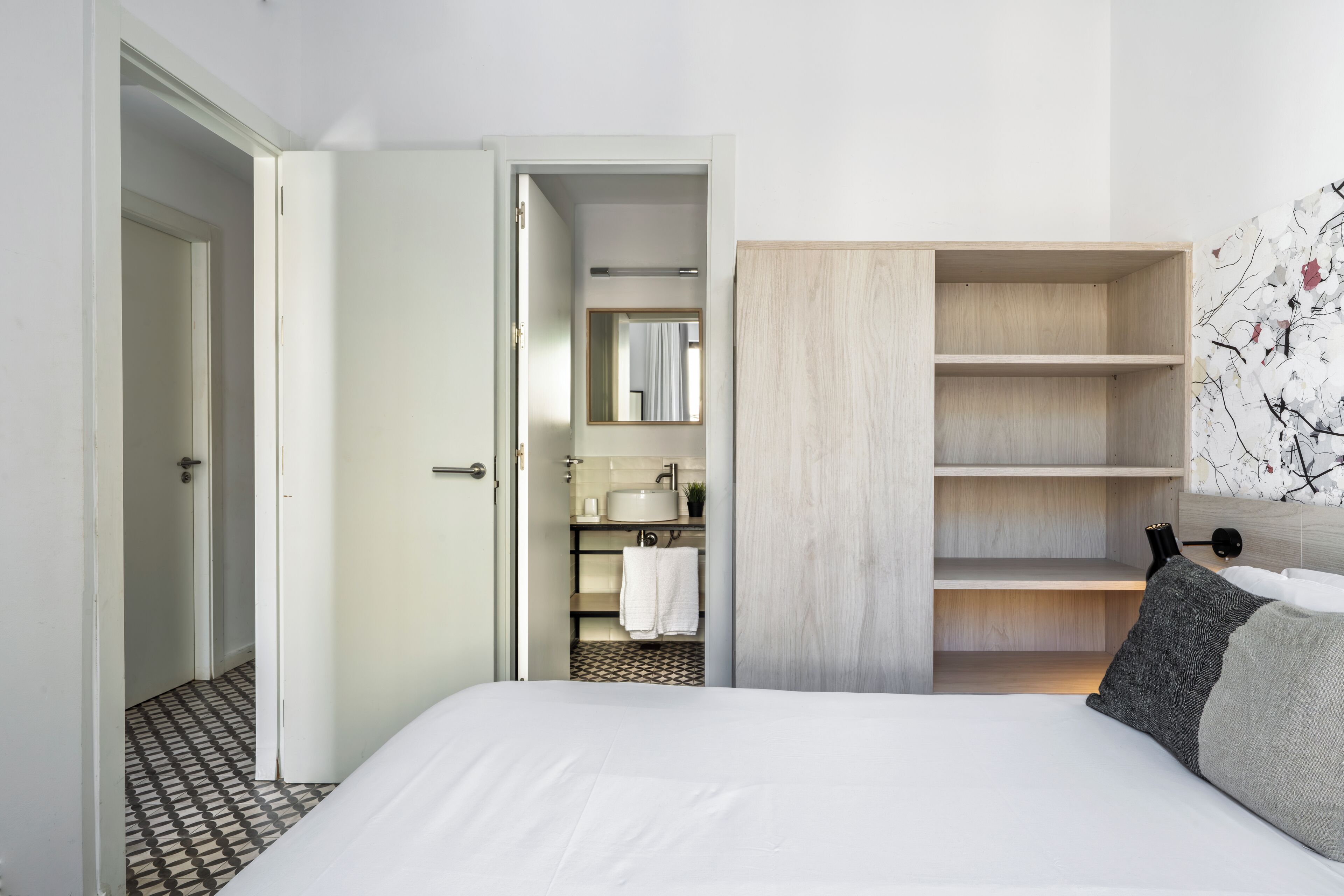 A modern hotel room with a neatly made bed, a wooden wardrobe, a bathroom with a sink and mirror, and a patterned floor.