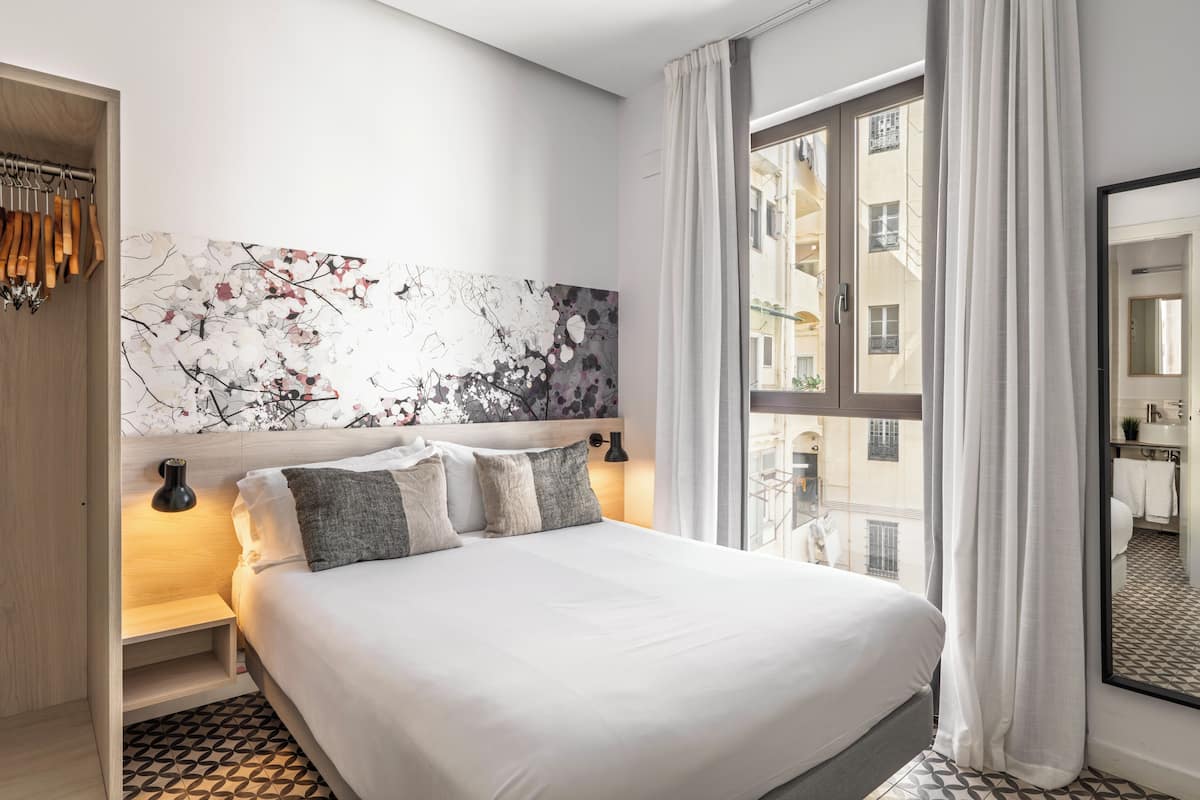 A modern hotel room with a large bed, a headboard with a floral artwork, a nightstand with a lamp, and a view of the city through the window.