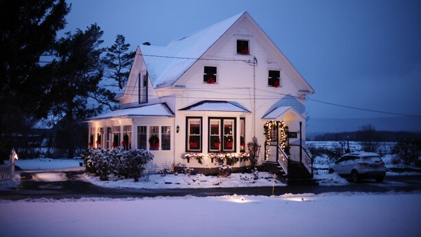 A large, white, two-story house with a covered porch, snow on the ground, and a car parked in front. - Carleton Inn and Cottages (Carleton Corner)