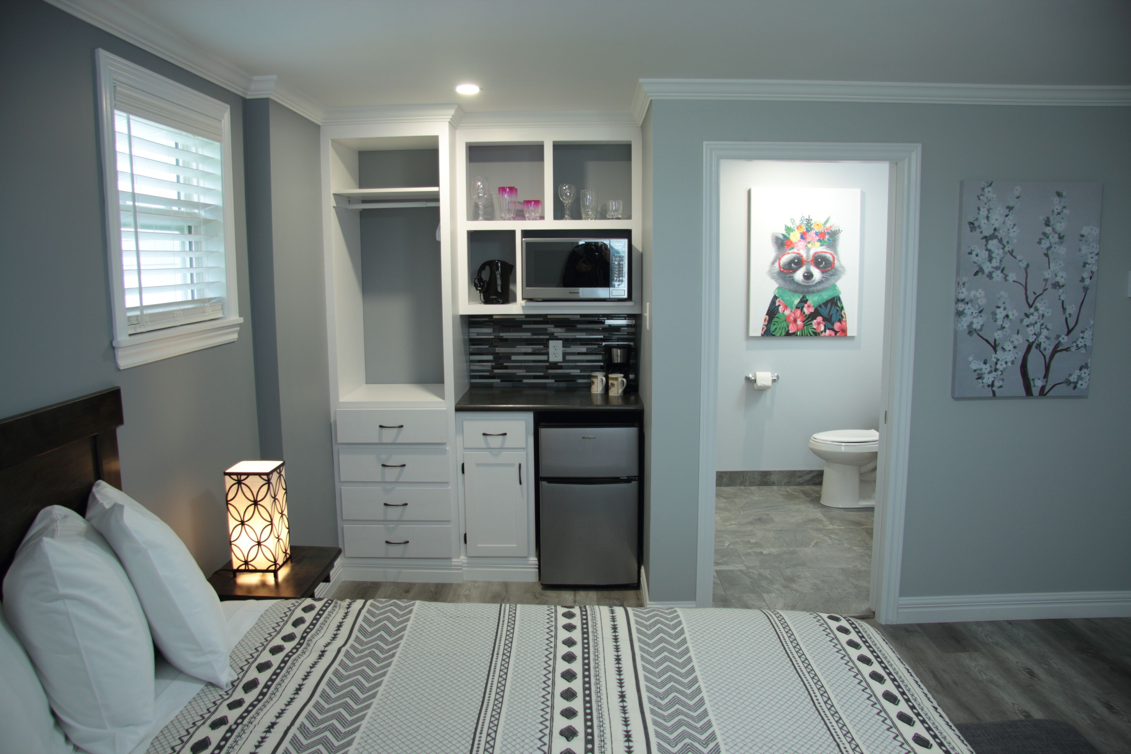 A bedroom with a bed, bedside lamp, and a view into a kitchen area with a refrigerator, microwave, and cabinets.