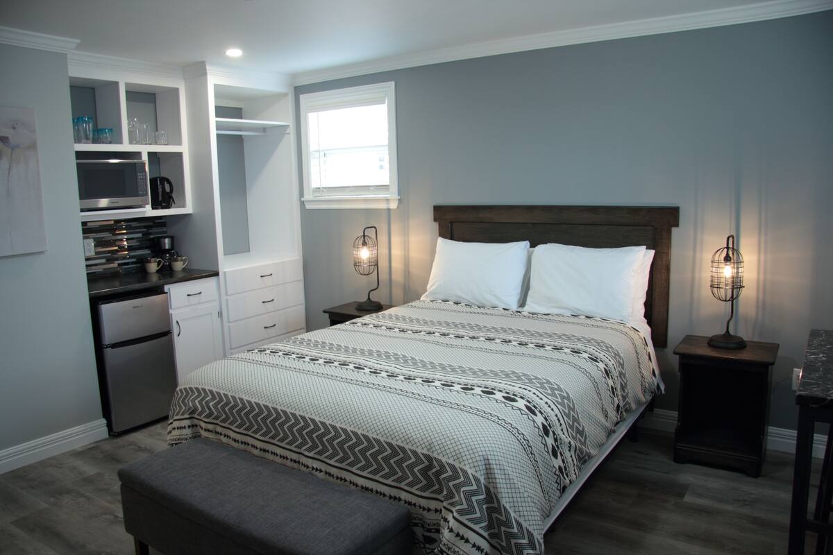 A bedroom with a bed, bedside tables, a bench, a built-in kitchenette, and a window.