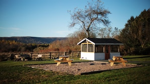A small white gazebo with a wooden roof and a door, surrounded by picnic tables and benches in a grassy area with trees and hills in the background. - Carleton Inn and Cottages (Carleton Corner)