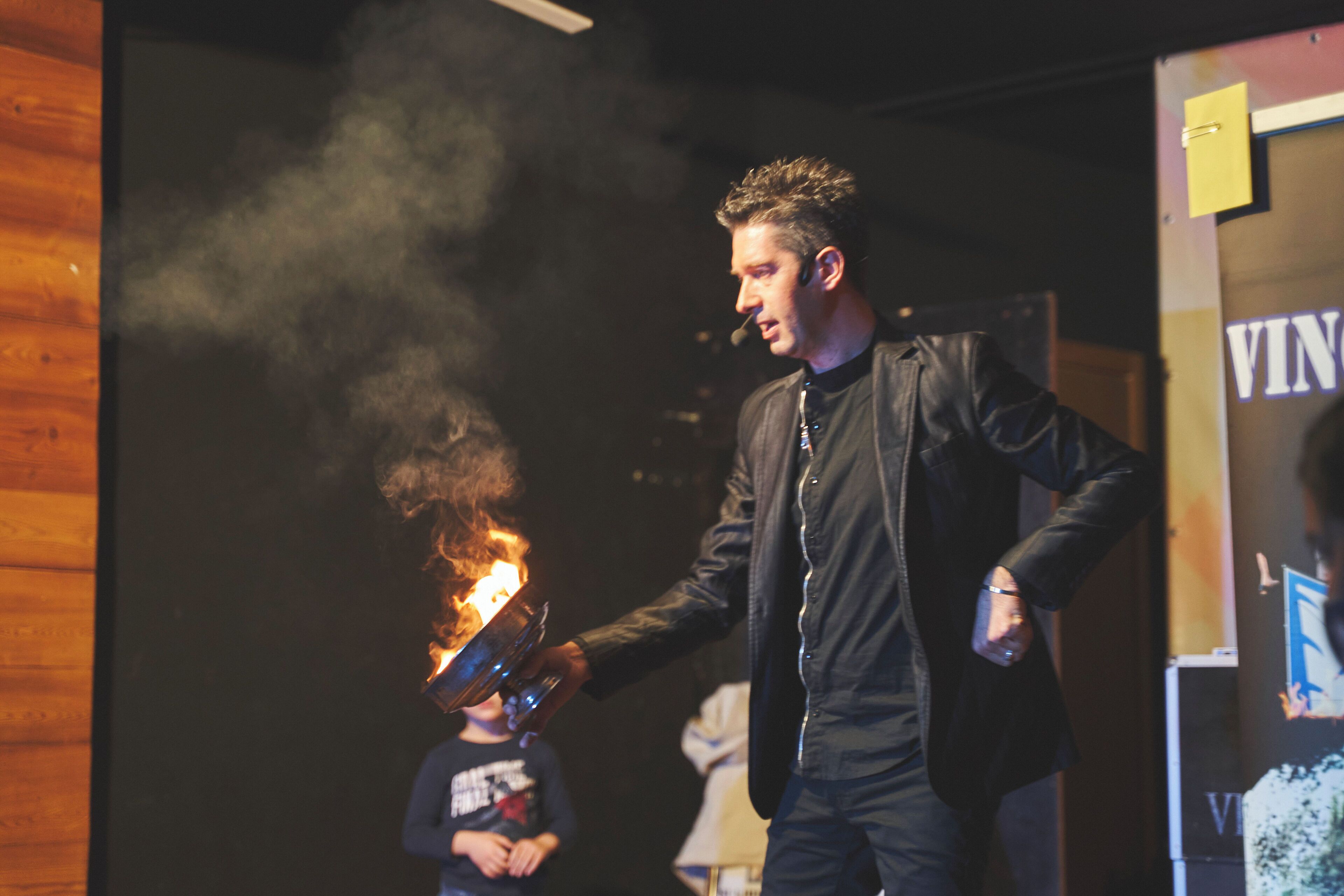A person holding a flaming object, with a wooden wall and a sign with the word "VIN" in the background.