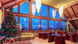 A cozy interior with a large Christmas tree, wooden furniture, and large windows offering a mountain view. - Hotel Mondolè (Frabosa Sottana)