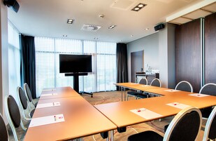 A conference room with a long table, chairs, a flat-screen TV, and a large window with blinds.