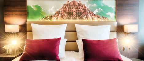 Habitación de hotel con una cama grande, dos almohadas, cabecera con un mural, lámparas de noche y una mesita con una lámpara.
