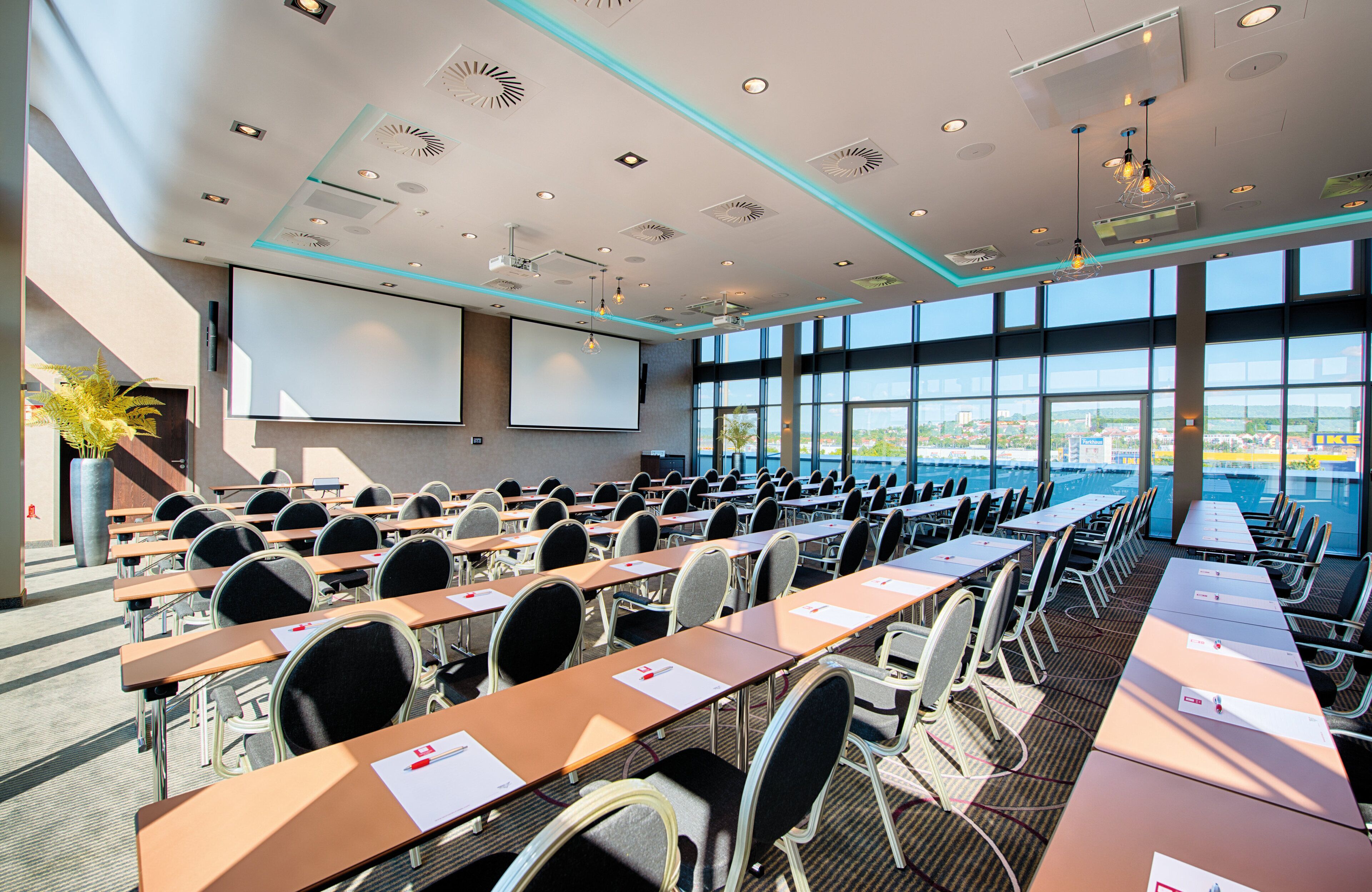A conference room with rows of tables and chairs, a large screen, and a view of the city through large windows.