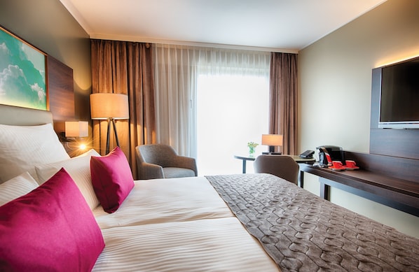 A hotel room with a large bed, a desk with a chair, a TV, and a window with curtains. - Leonardo Royal Hotel Ulm (Ulm)