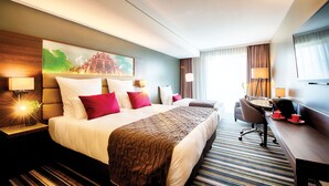 A hotel room with a large bed, a desk, a chair, and a painting on the wall. - Leonardo Royal Hotel Ulm (Ulm)
