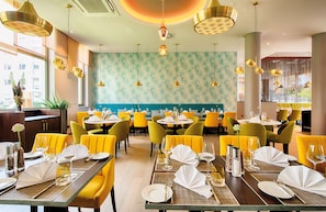 Un restaurante con sillas amarillas, mesas redondas y una pared azul turquesa estampada.