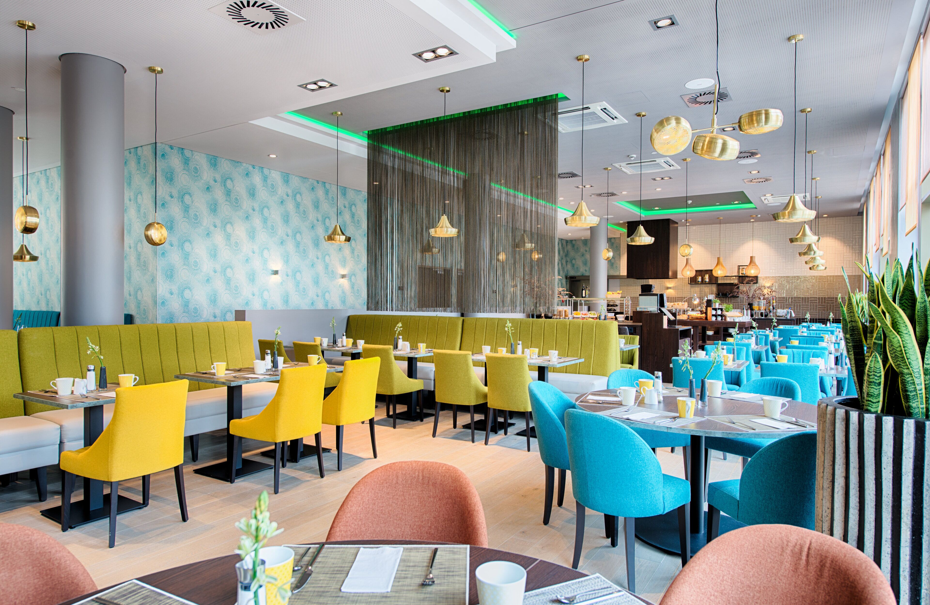 Ein modernes Restaurant mit Sitzboxen und Tischen, Polsterungen in Grün- und Gelbtönen und Pendelleuchten.