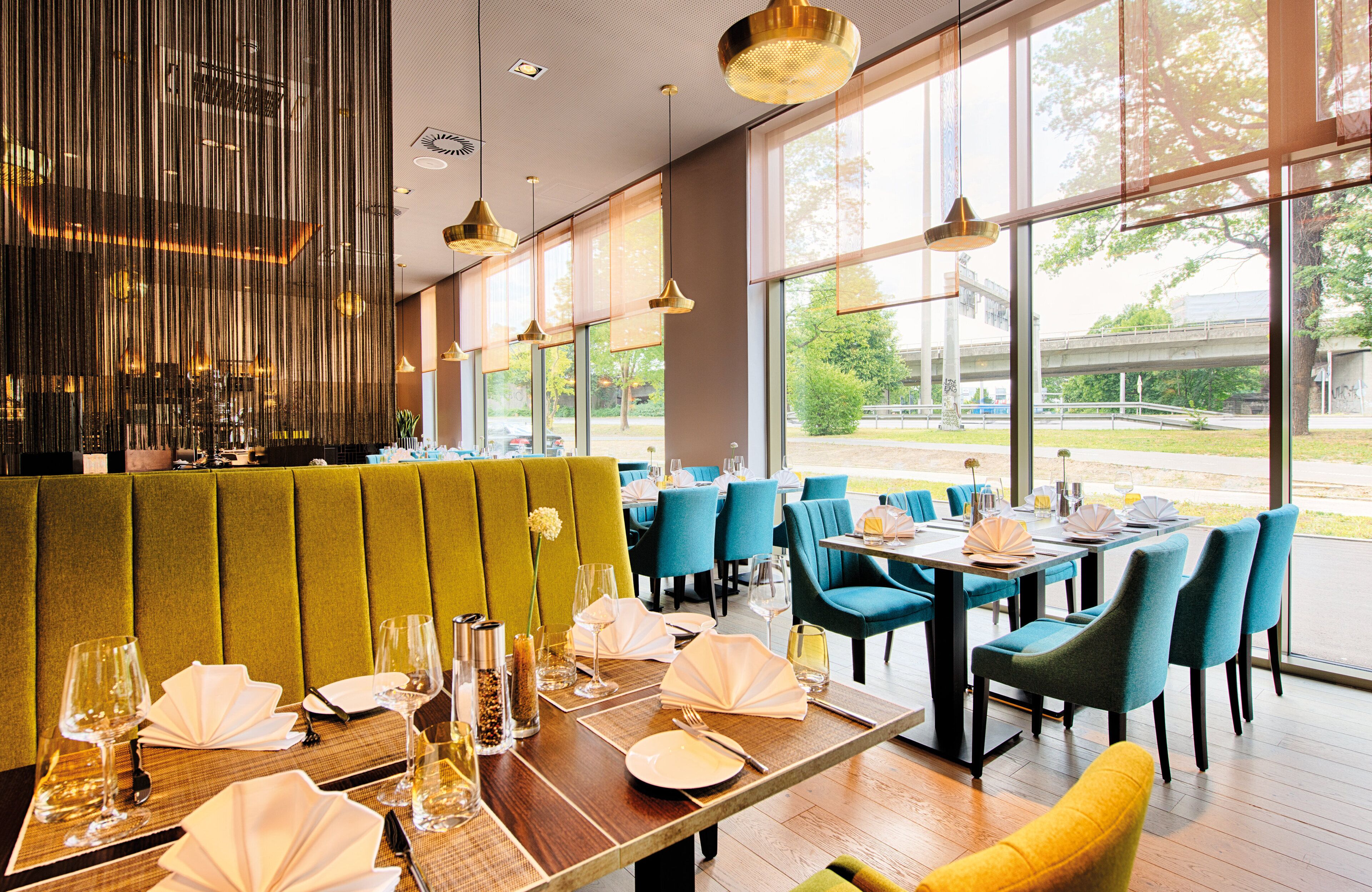 Ein Restaurant mit einer grünen Sitzgruppe, mehreren Tischen mit blauen Stühlen und großen Fenstern mit Blick ins Freie.