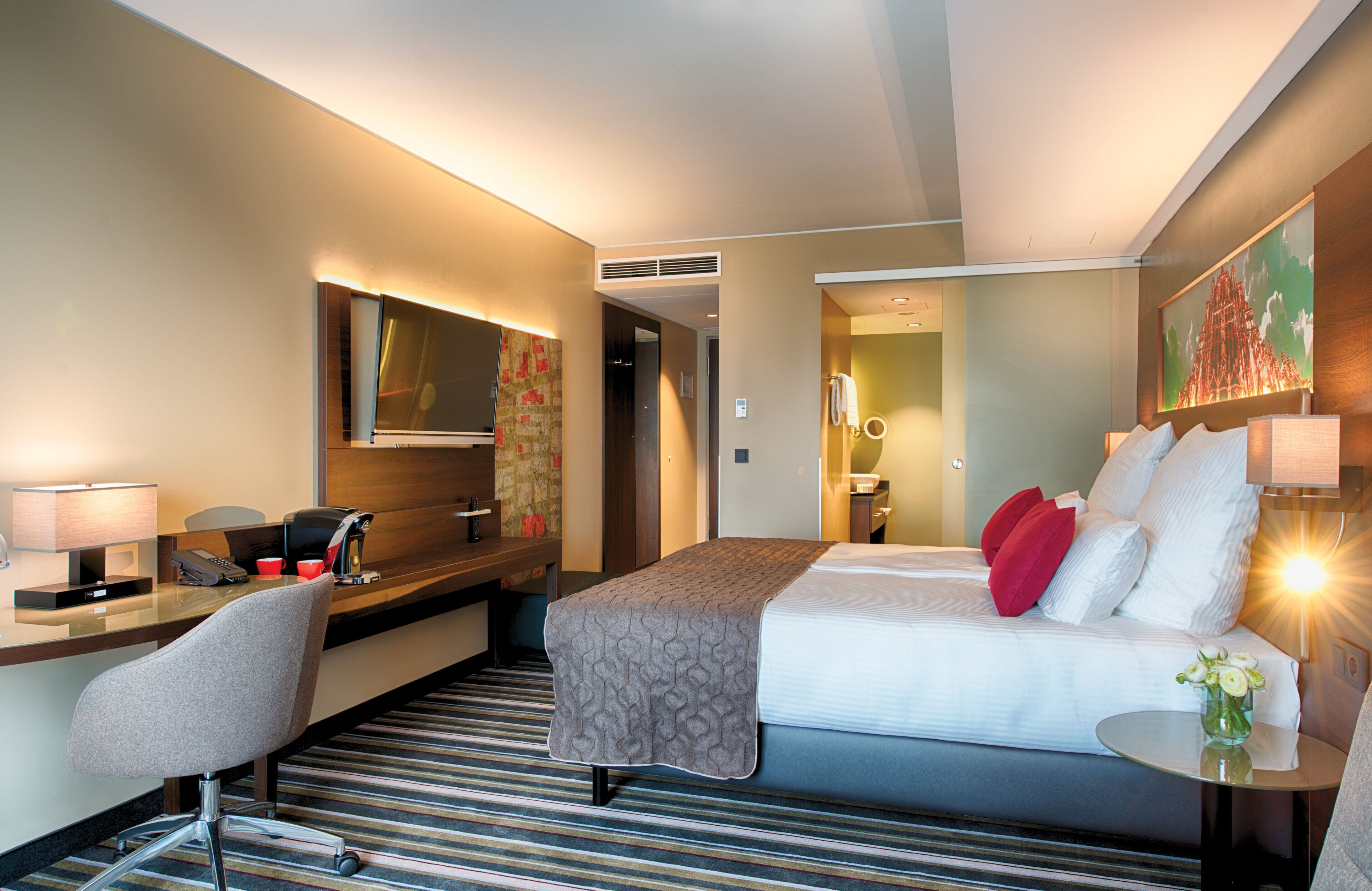 Habitación de hotel con cama, escritorio con teléfono y cafetera, una silla y una mesita con un florero.