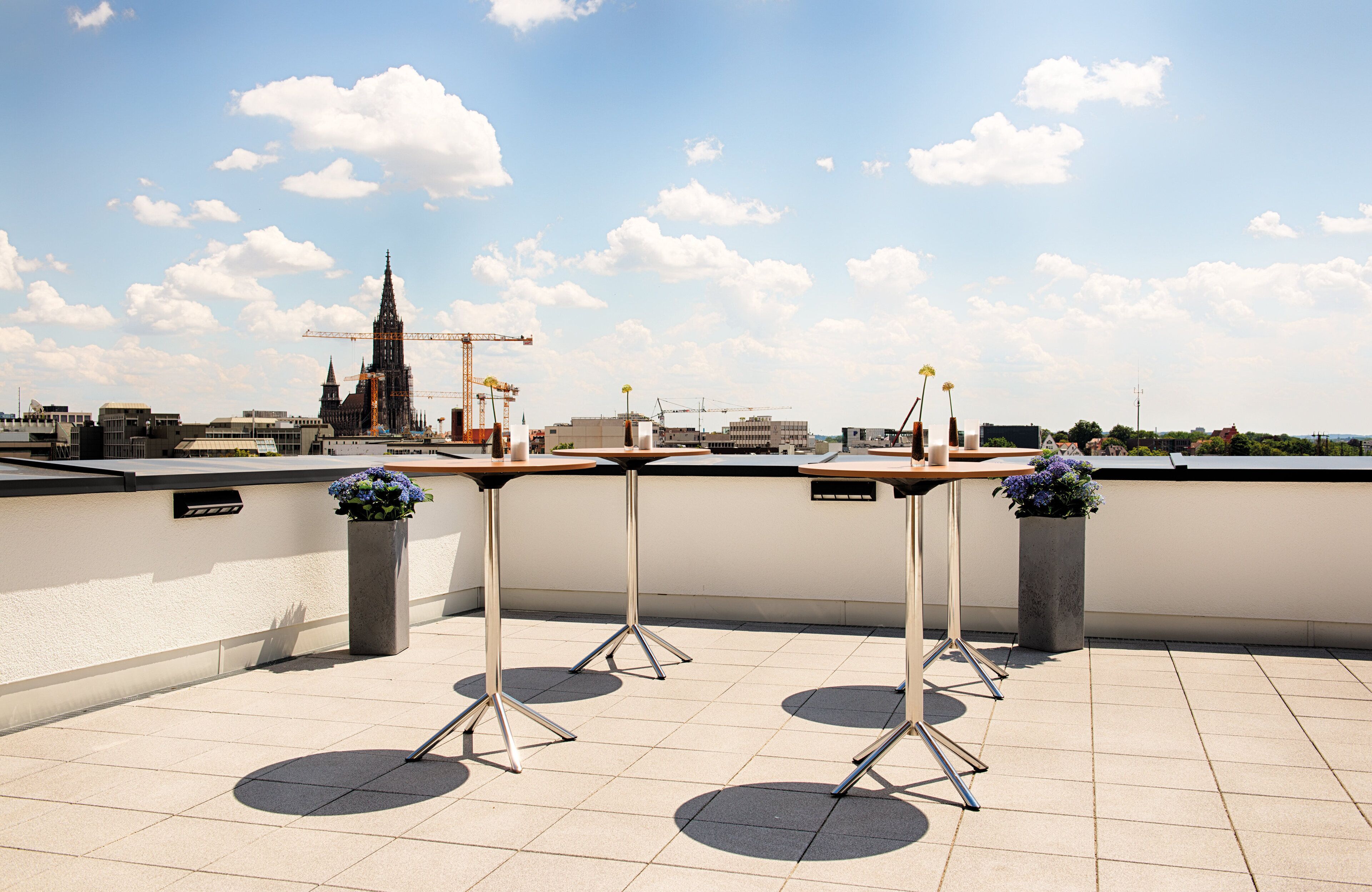 A rooftop terrace with tables and planters, overlooking a cityscape with a prominent cathedral and construction cranes.