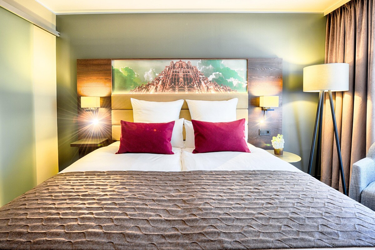 A hotel room with a large bed, two pillows, a headboard with a mural, bedside lamps, and a floor lamp.