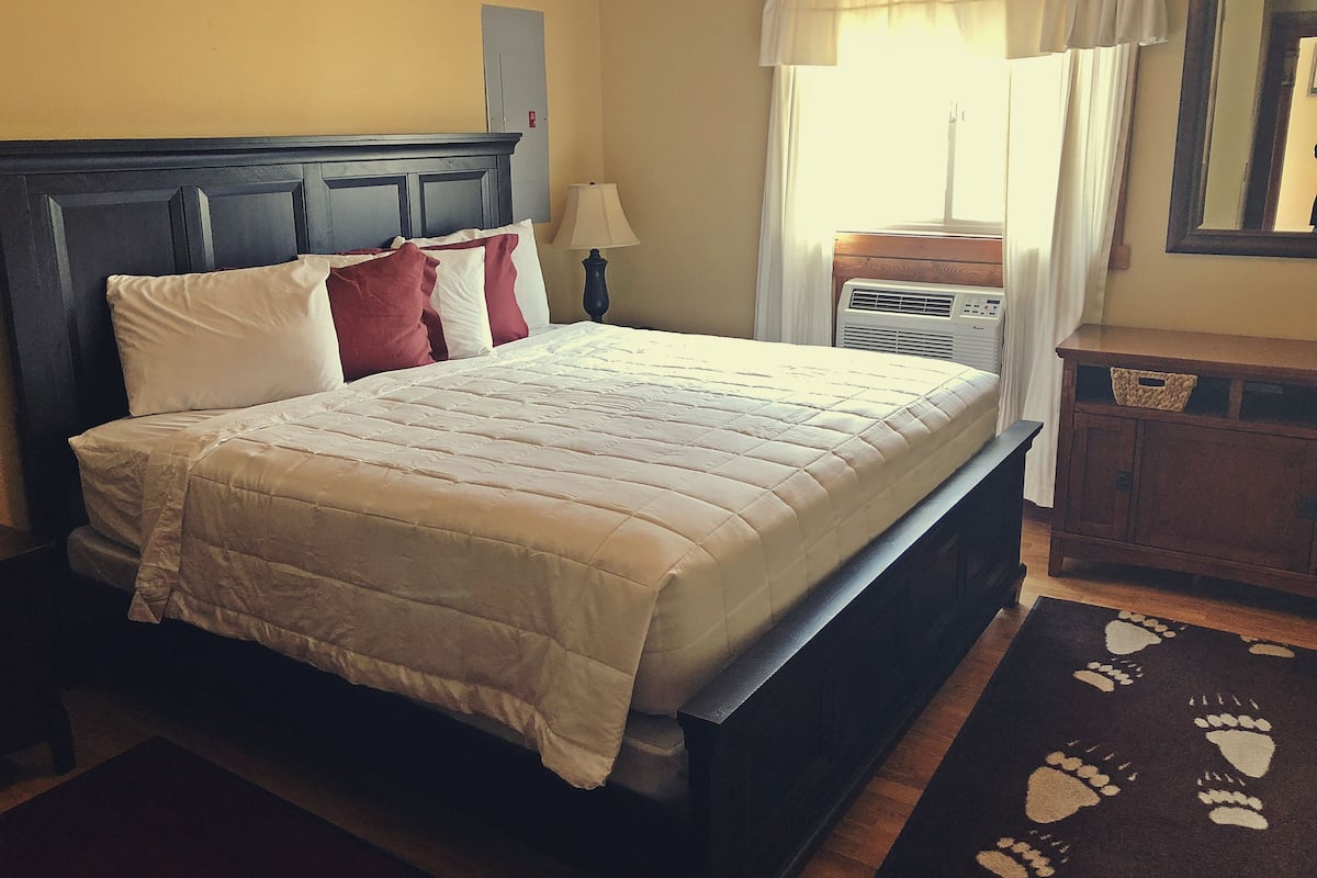 Una cama bien hecha con edredón blanco y almohadas rojas y blancas, cabecera de madera, lámpara de noche y mesita de noche de madera con una cesta.