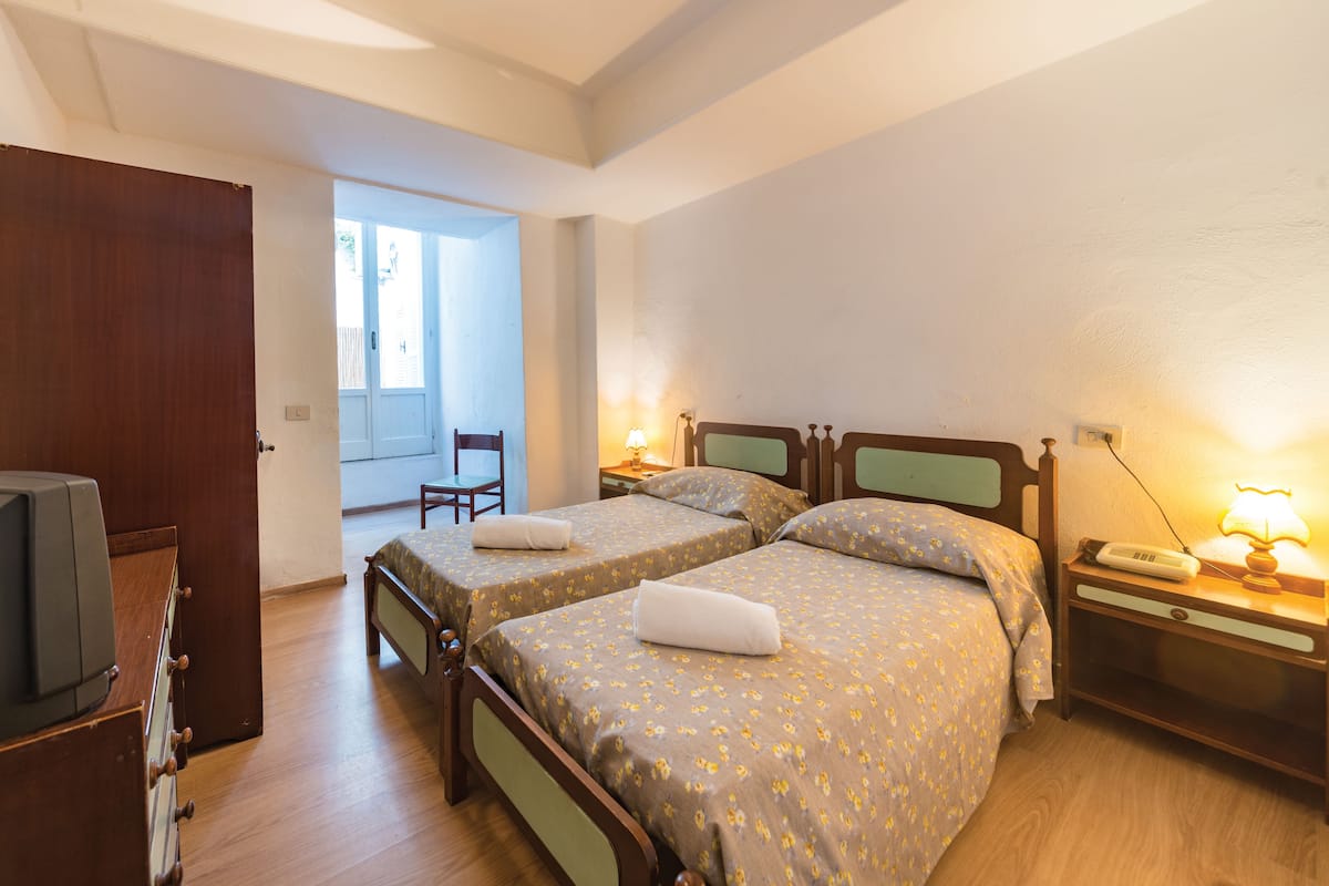 A hotel room with two single beds, wooden furniture, a desk with a lamp, and a television.
