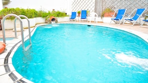 A swimming pool with blue water, surrounded by white tiles and blue lounge chairs. - Hotel Antonella (Ischia)
