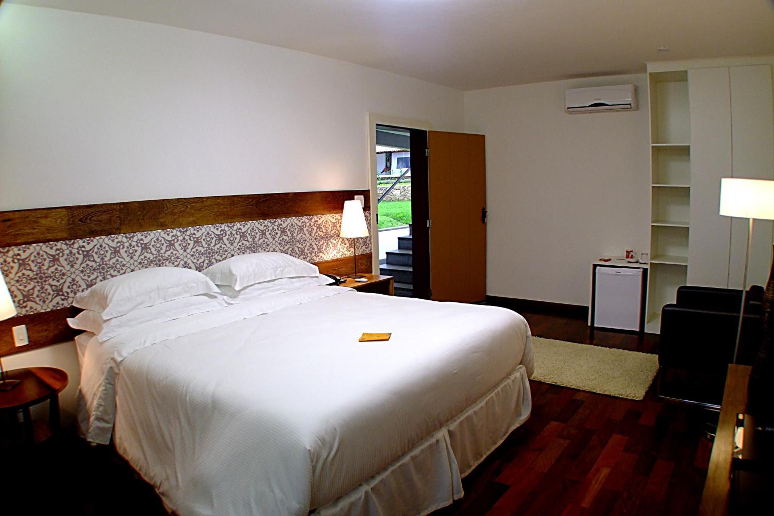 Een hotelkamer met een groot bed, nachtkastje, bureau, stoel, kleine tafel, lamp, boekenplank en een airconditioner.