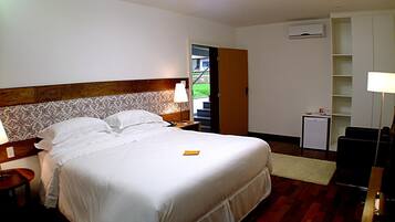 Een hotelkamer met een groot bed, nachtkastje, bureau, stoel, kleine tafel, lamp, boekenplank en een airconditioner.