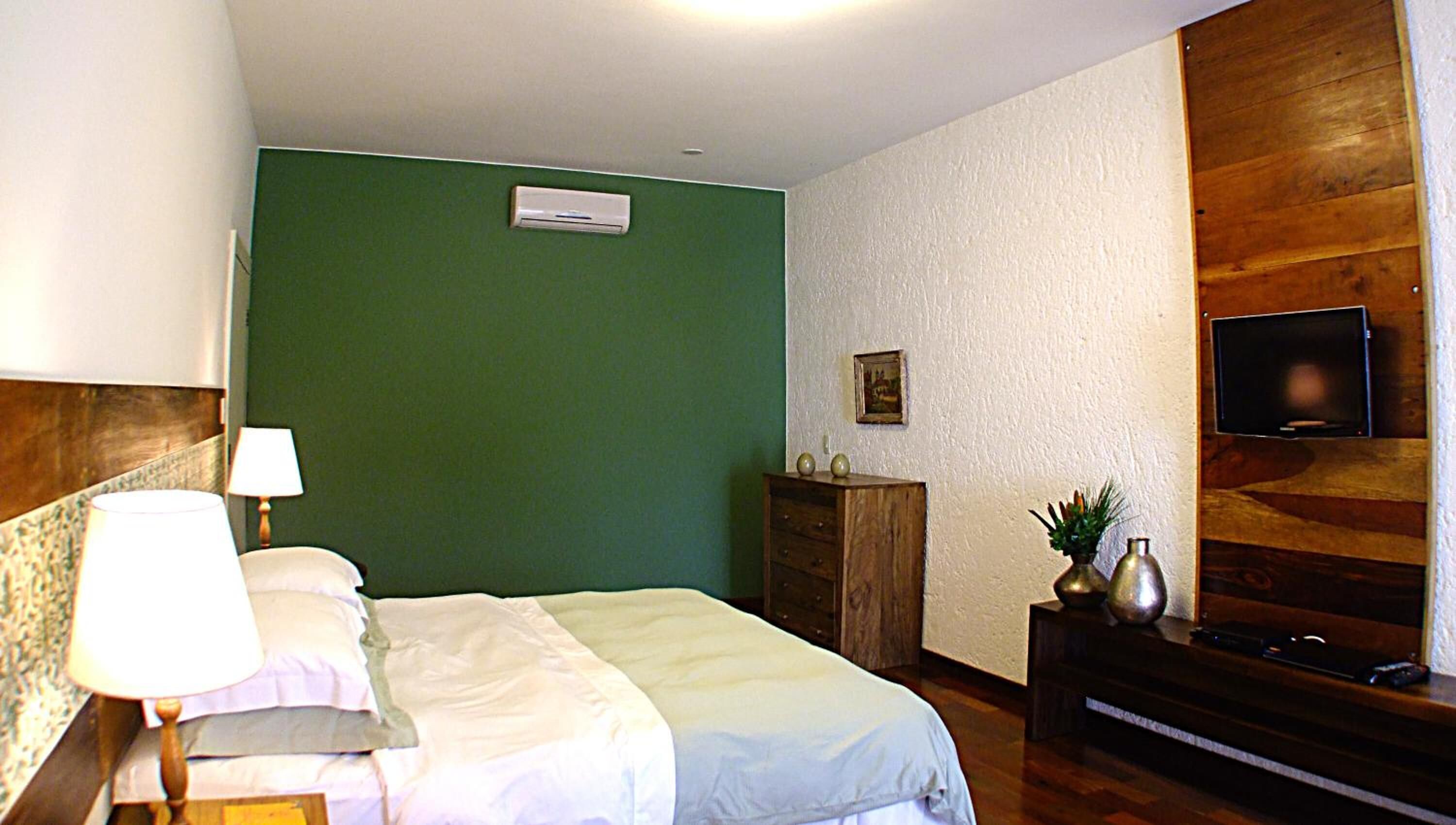 Une chambre d’hôtel avec un lit, une commode, un téléviseur fixé au mur et une petite table avec une lampe.