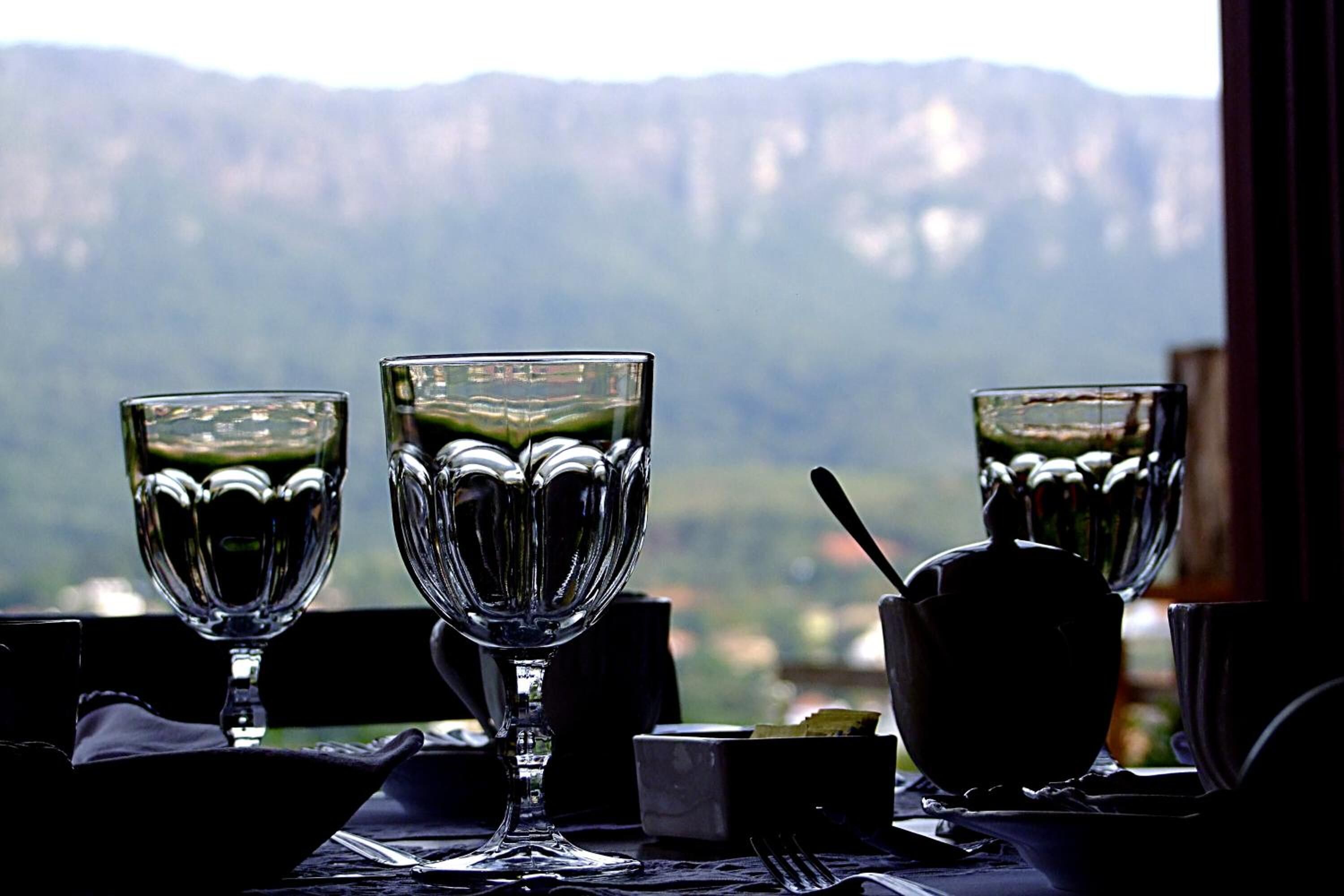 Una mesa con tres vasos con tallo y una tetera, frente a un paisaje montañoso.