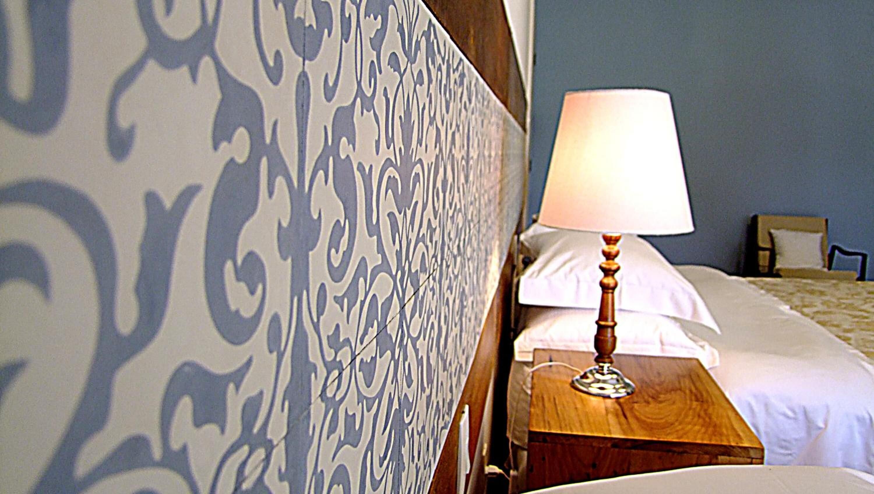 Una cama bien tendida con sábanas blancas, una mesita de noche de madera con una lámpara y un papel tapiz estampado.
