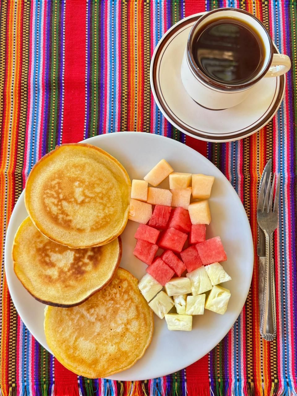 Une assiette de crêpes et de fruits, une tasse de café et une nappe colorée en tissu tissé.