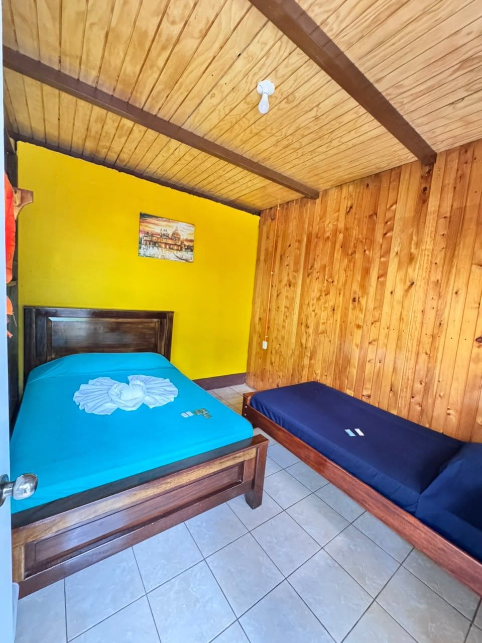 A room with a wooden ceiling, a bed with a blue bedspread, a wooden wall, and a small table.